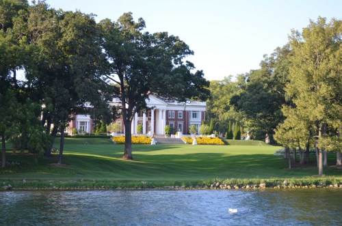 A large, elegant mansion surrounded by lush greenery and colorful flowers, viewed from across a calm lake.