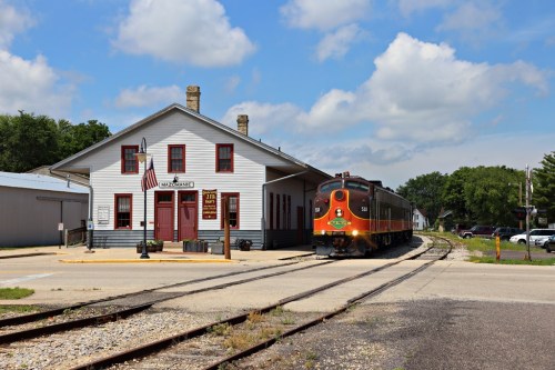 A vintage train approaches a historic train station under a blue sky with fluffy clouds.