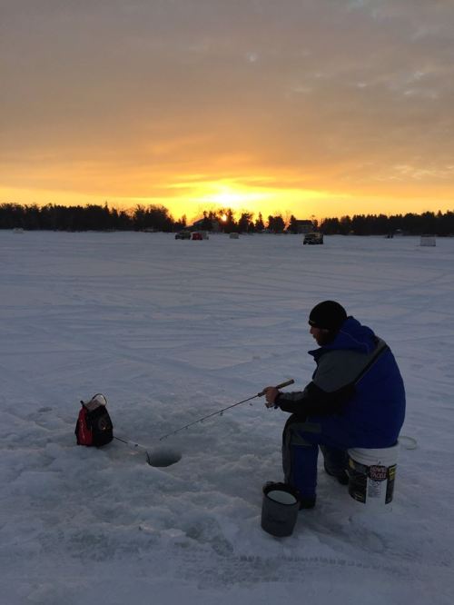 A person ice fishing on a snowy lake at sunset, with fishing gear and a hole in the ice.