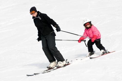 A person skiing with a child, who is being assisted on the slopes, both wearing winter gear and goggles.