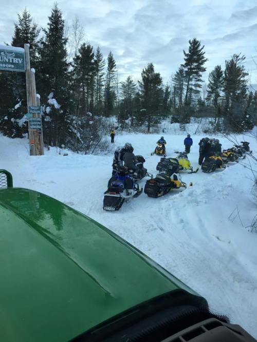 A snowy landscape with snowmobiles parked near a sign, and people gathered in the background among trees.