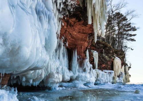 Icicles and frozen formations cling to a rocky cliff by a frozen lake, surrounded by trees under a clear sky.