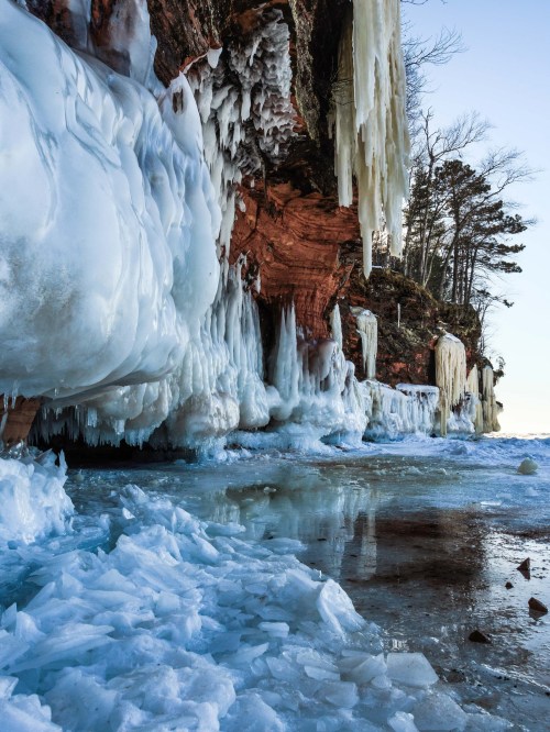Icicles hang from a rocky cliff, reflecting in icy water below, surrounded by a winter landscape.