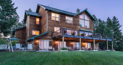 A large wooden house with multiple levels, surrounded by trees, featuring a spacious deck and large windows.