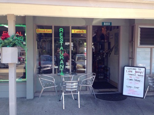 A small restaurant entrance with neon signs for breakfast and lunch, featuring outdoor seating and a menu board.