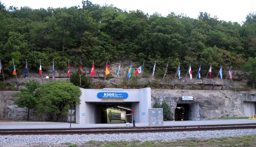 Entrance to a tunnel with flags from various countries displayed on a hillside above, near a railway track.