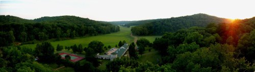 A panoramic view of a green valley with a building, tennis courts, and hills at sunset.
