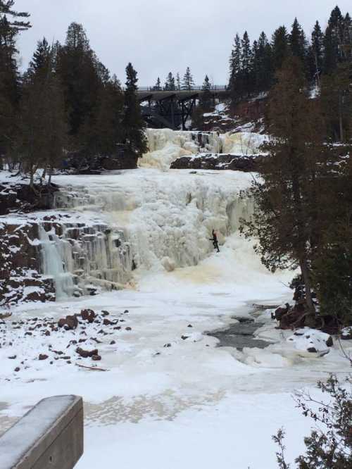 A frozen waterfall surrounded by snow-covered rocks and trees, with a bridge visible in the background.