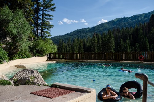 Two people relax in inner tubes in a natural pool surrounded by trees and mountains under a clear blue sky.