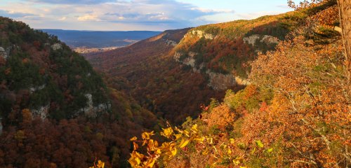 A scenic view of a valley surrounded by colorful autumn foliage and rocky cliffs under a partly cloudy sky.