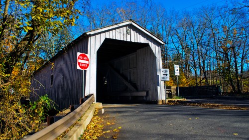 A white covered bridge with a wooden entrance, surrounded by autumn trees and road signs indicating no entry.