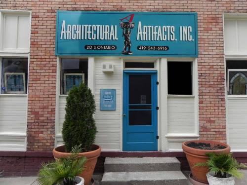 Front entrance of Architectural Artifacts, Inc. with a blue door and sign above, surrounded by potted plants.
