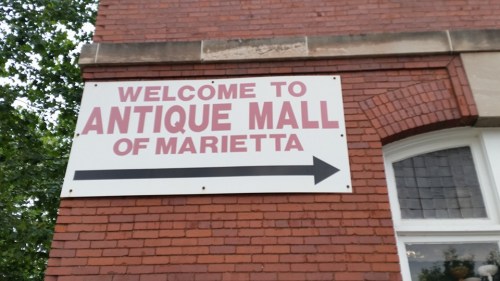 Sign on a brick wall reading "Welcome to Antique Mall of Marietta" with an arrow pointing to the left.