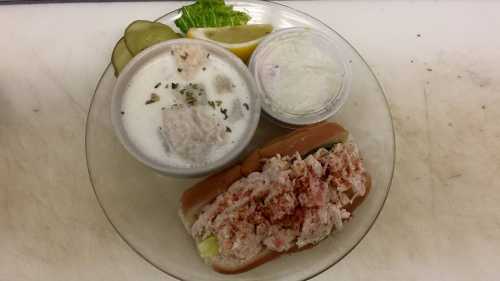 A plate featuring a lobster roll, a bowl of creamy soup, pickles, lettuce, and a lemon wedge.