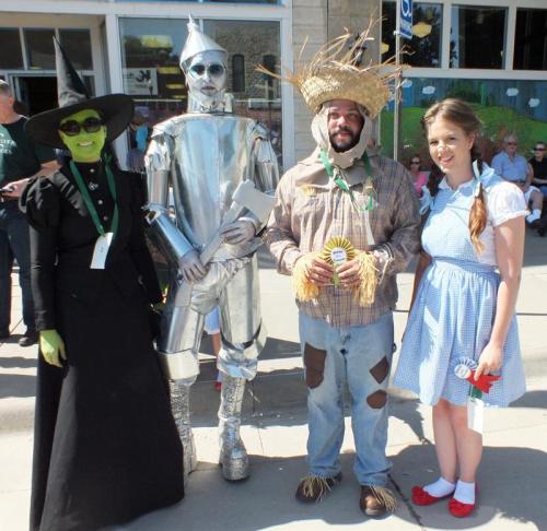Four people dressed as characters from "The Wizard of Oz": the Wicked Witch, Tin Man, Scarecrow, and Dorothy, smiling together.