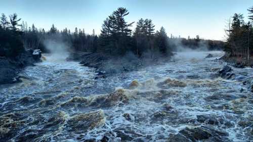 Raging river with frothy water flows through rocky terrain, surrounded by mist and evergreen trees in a serene landscape.