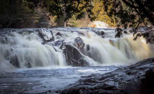 A serene waterfall cascades over rocky ledges, surrounded by lush greenery and soft sunlight.