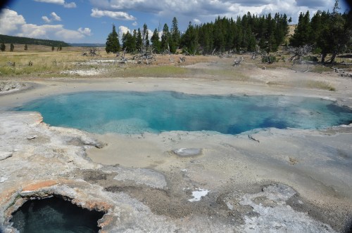 A vibrant blue hot spring surrounded by rocky terrain and green trees under a partly cloudy sky.