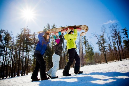 Three people carry a sled up a snowy hill under a bright sun, surrounded by trees.