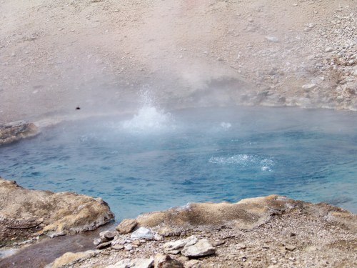 A steaming blue hot spring surrounded by rocky terrain, with water bubbling and splashing.