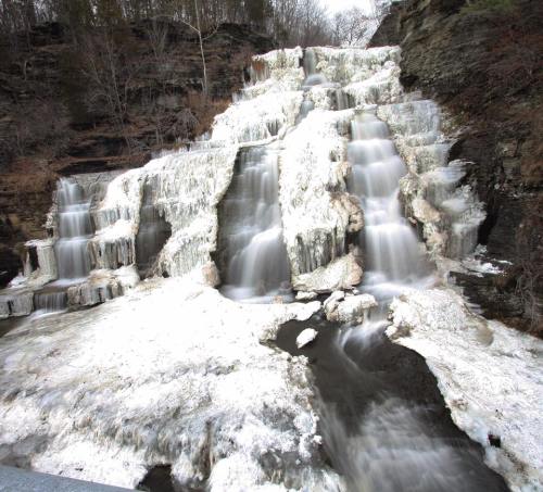 A frozen waterfall cascading down rocky cliffs, surrounded by ice and snow, creating a serene winter landscape.