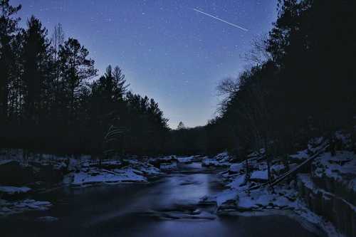 A serene night scene of a river surrounded by trees, under a starry sky with a shooting star.