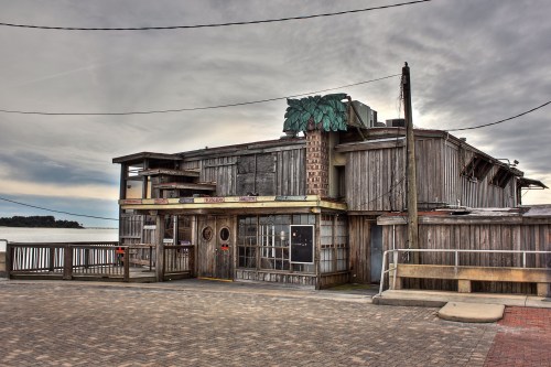 A weathered wooden building with a palm tree sign, overlooking a calm body of water under a cloudy sky.