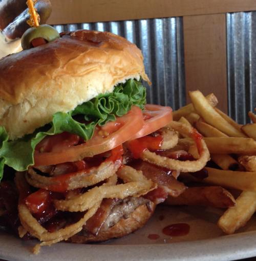 A large burger with lettuce, tomato, bacon, and crispy onions, served with a side of golden fries.