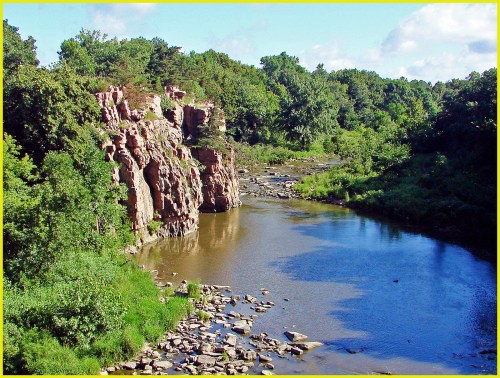 A serene river flows past rocky cliffs and lush greenery under a clear blue sky.
