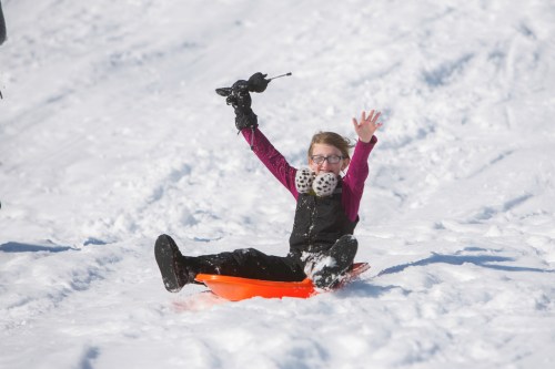 A child in winter gear joyfully sleds down a snowy hill, raising one hand in excitement.