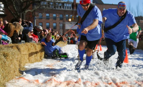 Two men in blue shirts race on snow while a crowd watches, with one man falling behind on a sled.