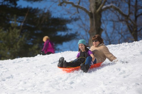 A woman and a girl joyfully sled down a snowy hill, with another person in the background enjoying the winter day.