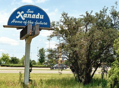 Sign for Xanadu, "Home of the Future," next to a tree and a road with a car passing by under a blue sky.