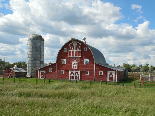 A red barn with a curved roof and a silo, surrounded by green grass and under a cloudy blue sky.