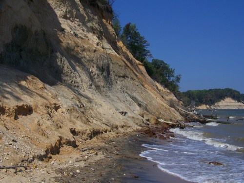 A sandy shoreline with steep cliffs, trees in the background, and gentle waves lapping at the beach under a clear blue sky.