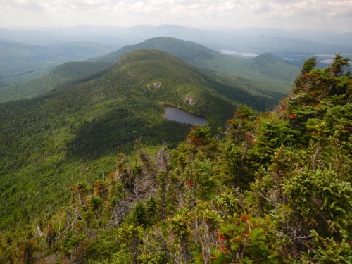 A panoramic view of lush green mountains and a small lake nestled in the valley under a cloudy sky.