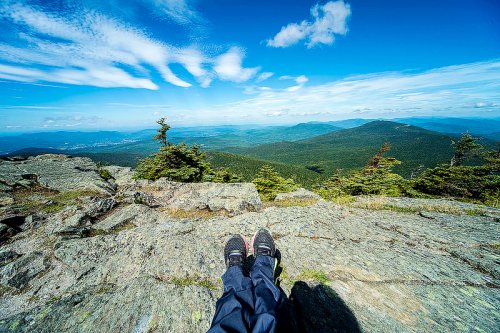 Person's feet resting on a rocky ledge, overlooking a vast green landscape and blue sky with scattered clouds.