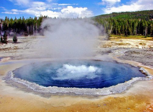 A vibrant blue hot spring surrounded by steam and a rocky edge, set against a backdrop of green trees and blue sky.
