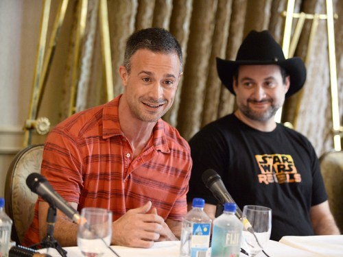 Two men sit at a table with microphones, one in a red striped shirt and the other in a black cowboy hat and t-shirt.