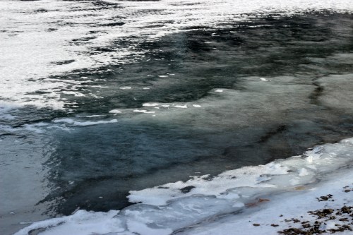 A partially frozen lake with dark water and patches of ice along the shore, reflecting a cloudy sky.
