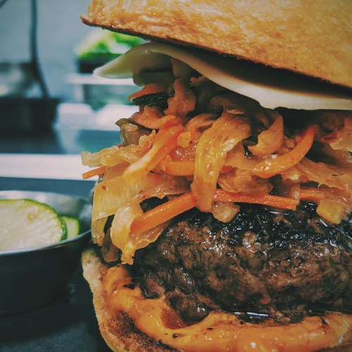 A close-up of a juicy burger topped with sautéed onions and sauce, served with pickles on the side.