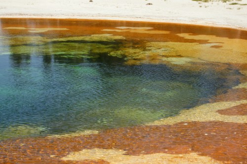 A colorful hot spring with vibrant orange and green hues, surrounded by sandy terrain.
