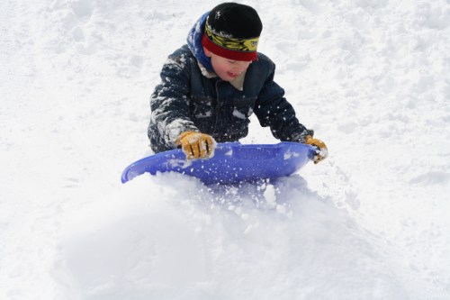A child in winter clothing sleds down a snowy hill, joyfully kicking up snow.