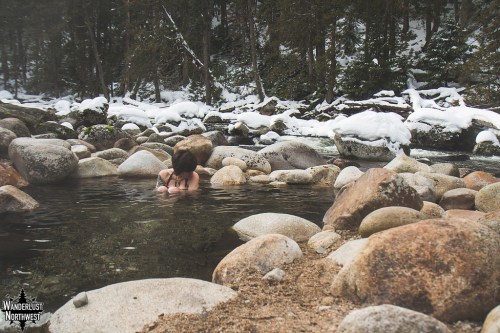 A person sits in a natural pool surrounded by large rocks and snow-covered trees, enjoying a serene outdoor setting.