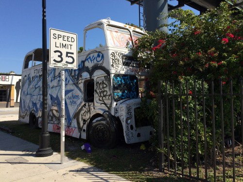 A graffiti-covered bus partially hidden by bushes, next to a speed limit sign in an urban setting.