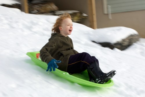 A child joyfully sledding down a snowy hill, wearing a green jacket and blue gloves, with hair blowing in the wind.