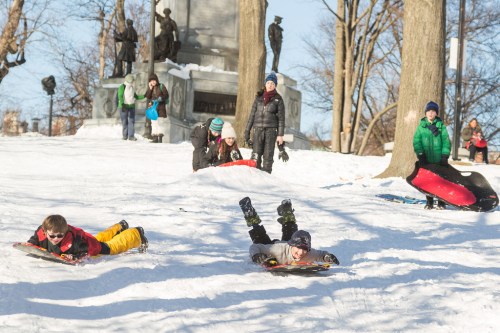 Children sledding down a snowy hill, with adults watching and a monument in the background.
