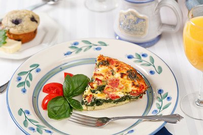 A slice of vegetable frittata on a decorative plate, garnished with cherry tomatoes and basil, alongside a muffin and drink.