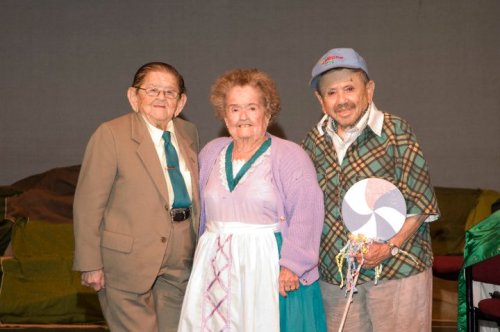 Three elderly individuals pose together on stage, smiling. One wears a suit, another a dress, and the third holds a lollipop.