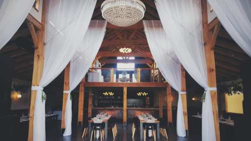 Elegant restaurant interior with wooden beams, a chandelier, and tables set for dining, draped in white curtains.